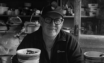 Man with trimmed white beard and cap. Interior with shelves.