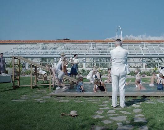 Back view of White man with shaven head in white suit standing looking at a swimming pool full of children, sunny day